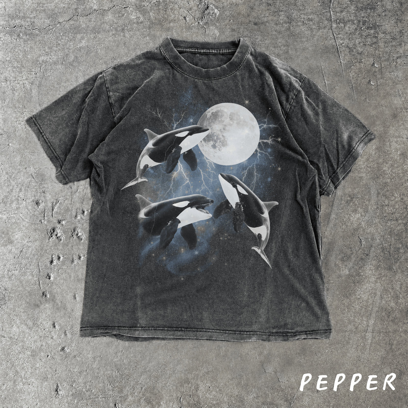A washed black graphic t-shirt featuring three orcas swimming around a full moon, laid flat on a textured gray concrete background. The word 'PEPPER' is printed in white in the bottom right corner.